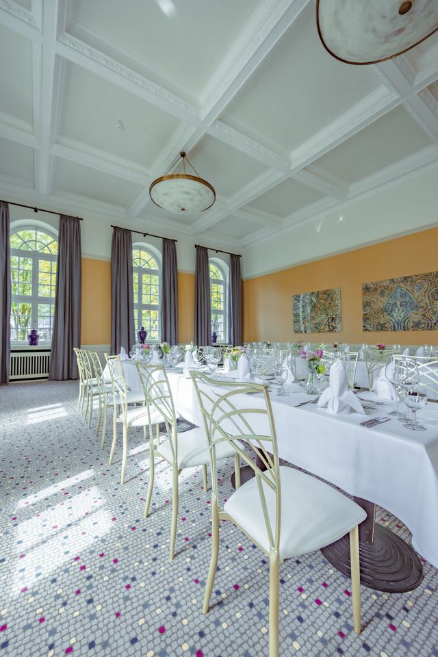 Ein Esszimmer, gedeckt für ein Bankett. Weiße Stühle und Tische auf gemustertem Teppich, Bogenfenster, dekorative Decke.