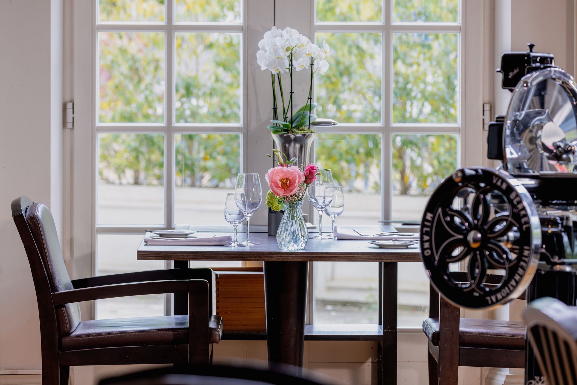 Ein gedeckter Tisch mit frischen Blumen vor einem hellen Fenster, daneben eine Fleischschneidemaschine im Vintage-Stil.