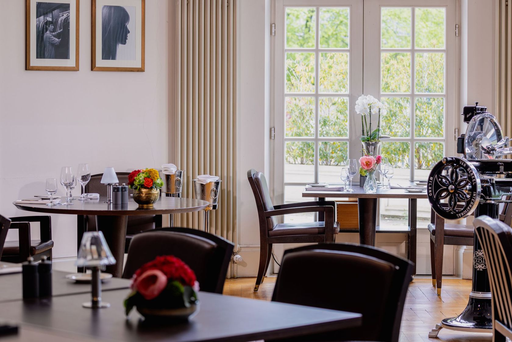 Restaurant-Speisesaal mit Tischen, Stühlen und großen Fenstern mit Blick ins Grüne. Blumengestecke schmücken die Tische.