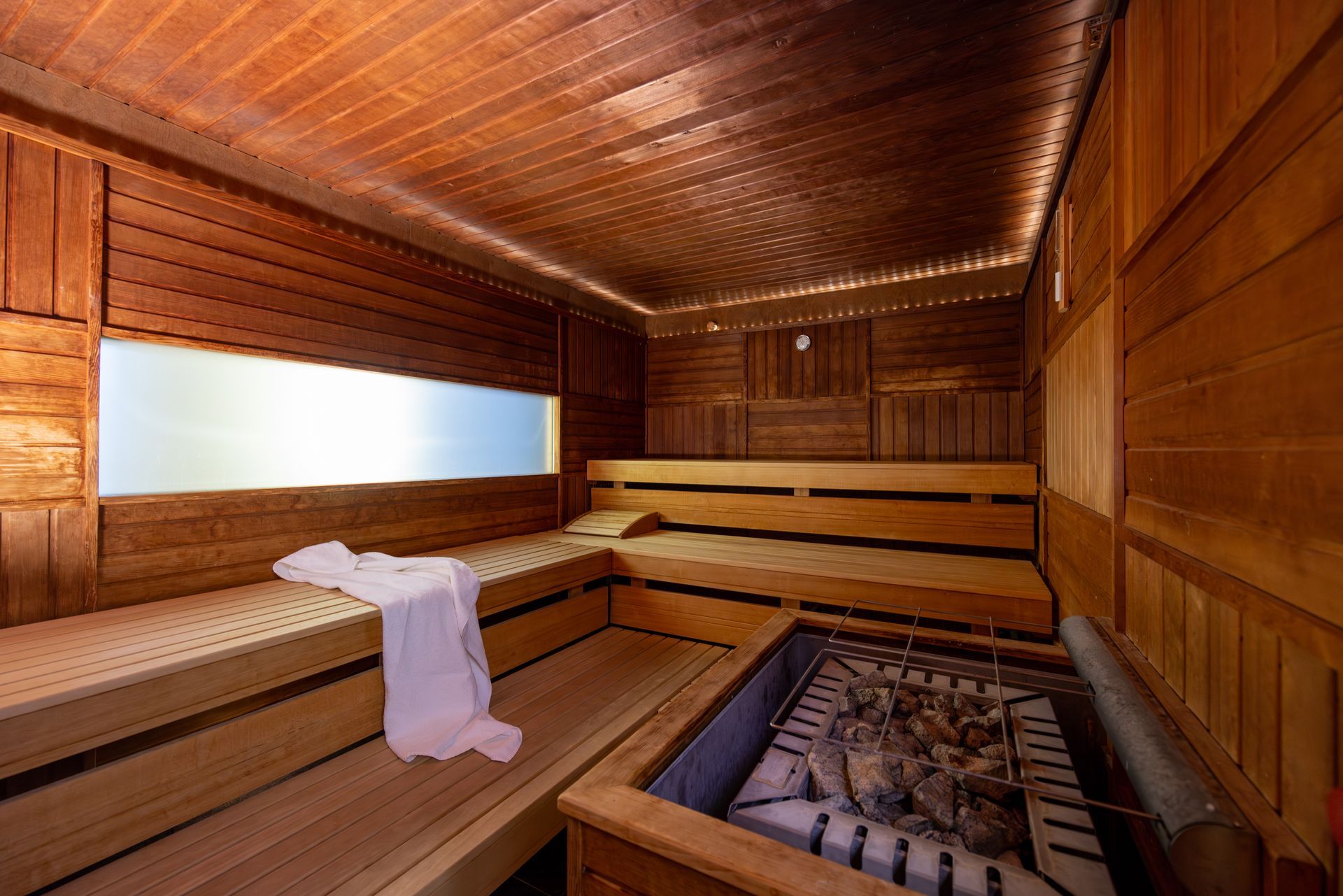 Die Sauna ist aus Holz gefertigt und mit Bänken, einer Heizeinheit und einem Fenster ausgestattet, durch das Licht hereinfällt. Auf einer Bank liegt ein weißes Handtuch.