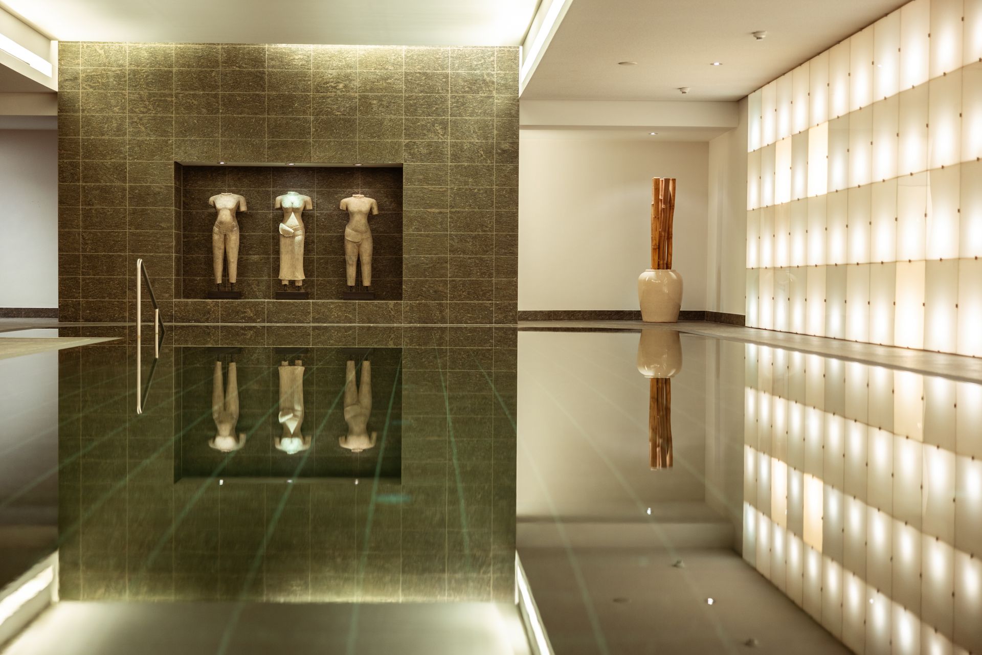 Ein reflektierender Teich mit Skulpturen, beleuchteten Wandpaneelen und einem vertikalen Holzkunstwerk in einem modernen Ambiente.