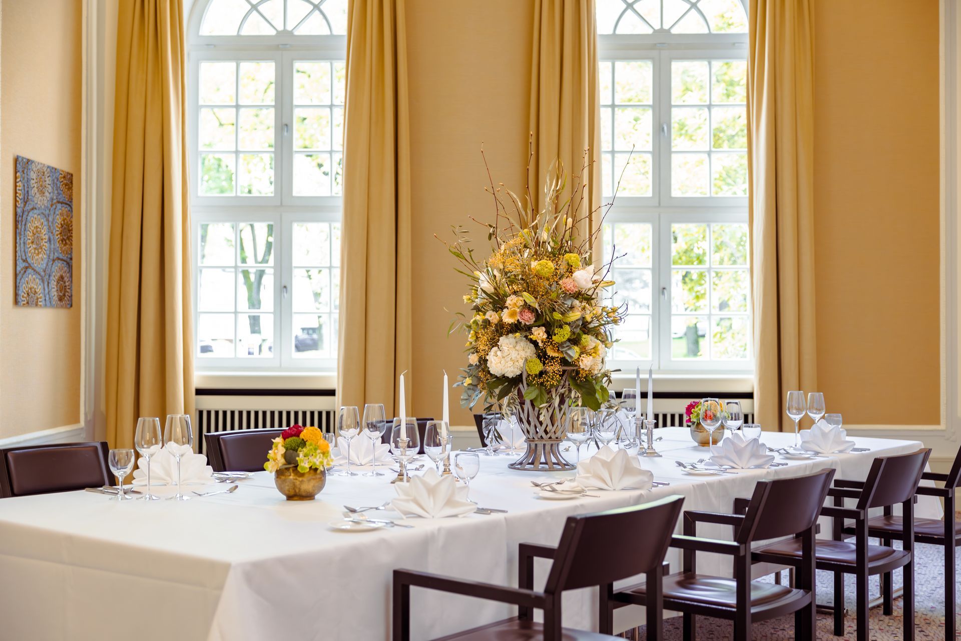 Festlich gedeckter Speisesaal für eine Feier. Weiße Tischdecke, Blumengesteck, Stühle und Fenster mit goldenen Vorhängen.