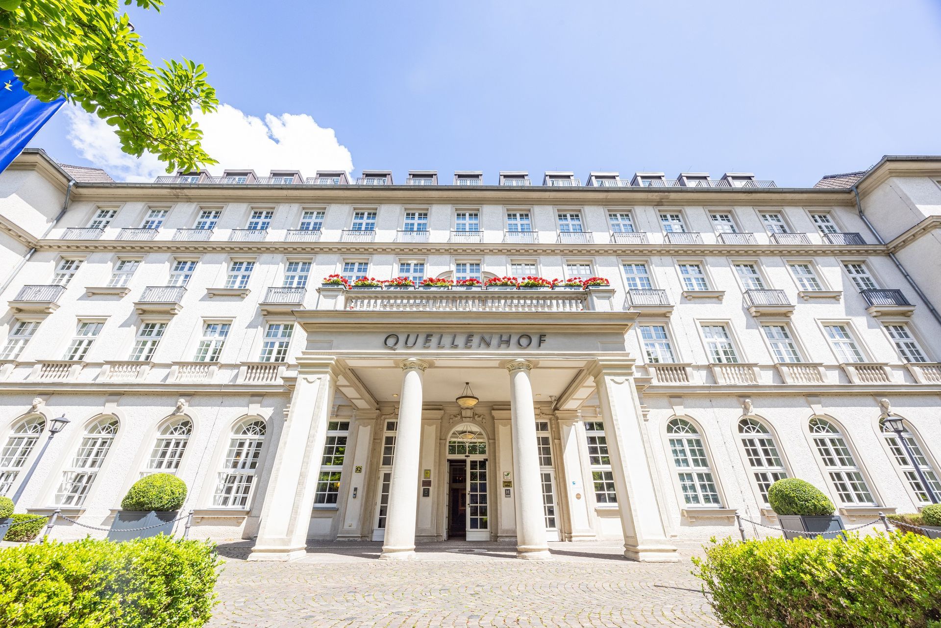 Une chambre d'hôtel avec deux fauteuils verts, une table d'appoint, un lampadaire, un bureau et une télévision accrochée au mur.