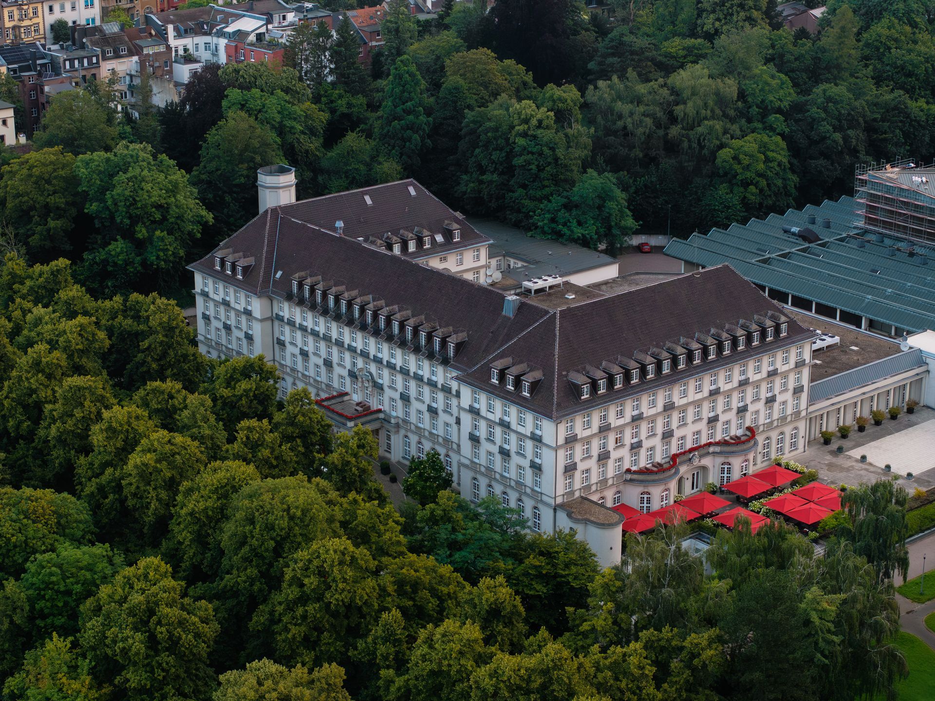 Großes weißes Gebäude mit dunklem Dach, umgeben von grünen Bäumen. Rote Markisen sind sichtbar.