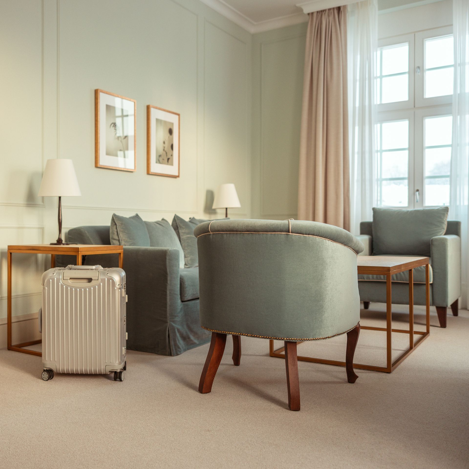 Ein Hotelzimmer mit hellgrünen Wänden, einer Couch, Stühlen, Lampen, Kunstwerken, einem Koffer und einem Fenster mit einem durchsichtigen Vorhang.