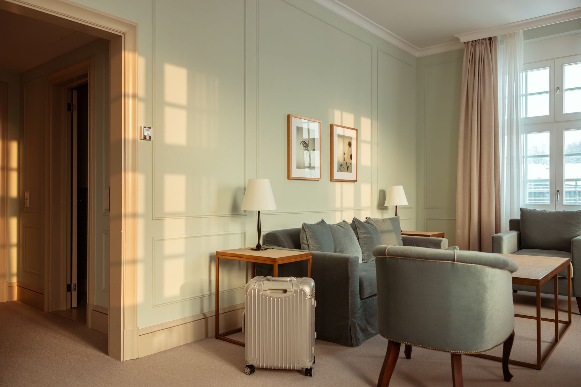 Living room with a sofa, armchairs, side tables, a window, and luggage. Sunlight streams in.