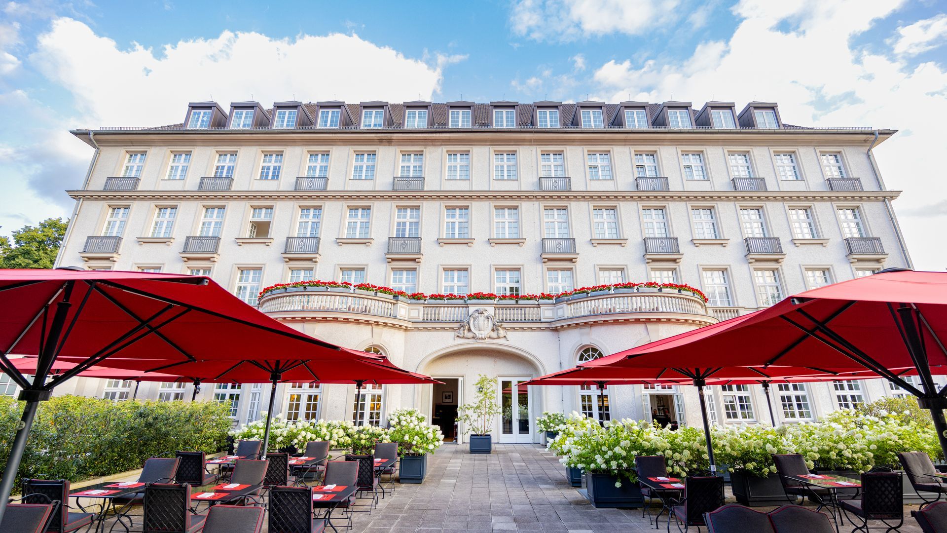 Hotelfassade mit roten Sonnenschirmen, die die Außensitzplätze beschatten. Cremefarbenes Gebäude mit vielen Fenstern und Blumenschmuck.
