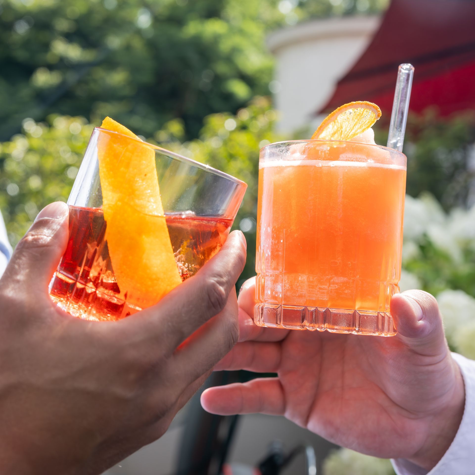Zwei Hände halten Cocktails: in der einen eine Orangenschale, in der anderen eine Orangenscheibe. Draußen. Prost!