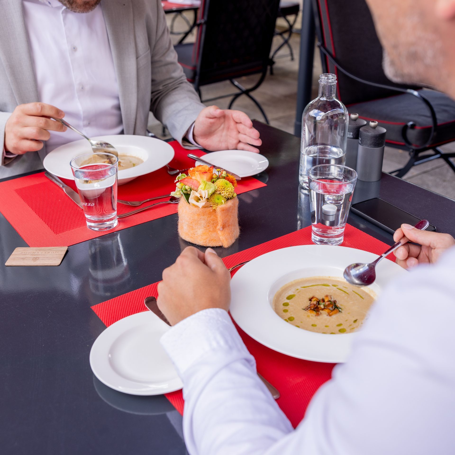 Zwei Personen essen Suppe an einem Restauranttisch; rote Platzsets, Blumen, Wasserflaschen.