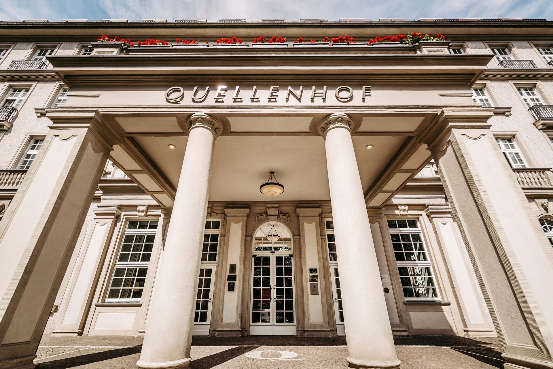 Hoteleingang mit großen Säulen und dem Schriftzug „Quellenhof“ über der Tür. Weißes Gebäude mit vielen Fenstern, blauer Himmel.