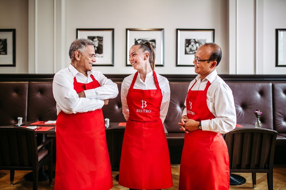 Three people wearing red aprons and white shirts are standing in a restaurant, smiling.