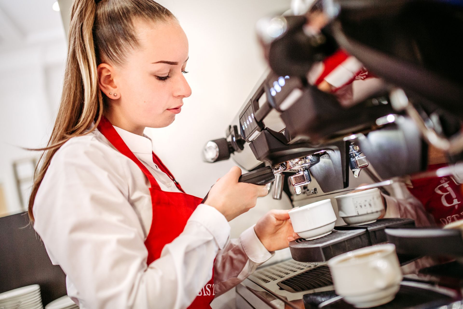 A barista is making espresso; he is wearing a red apron and a white shirt and is holding a cup in his hand.
