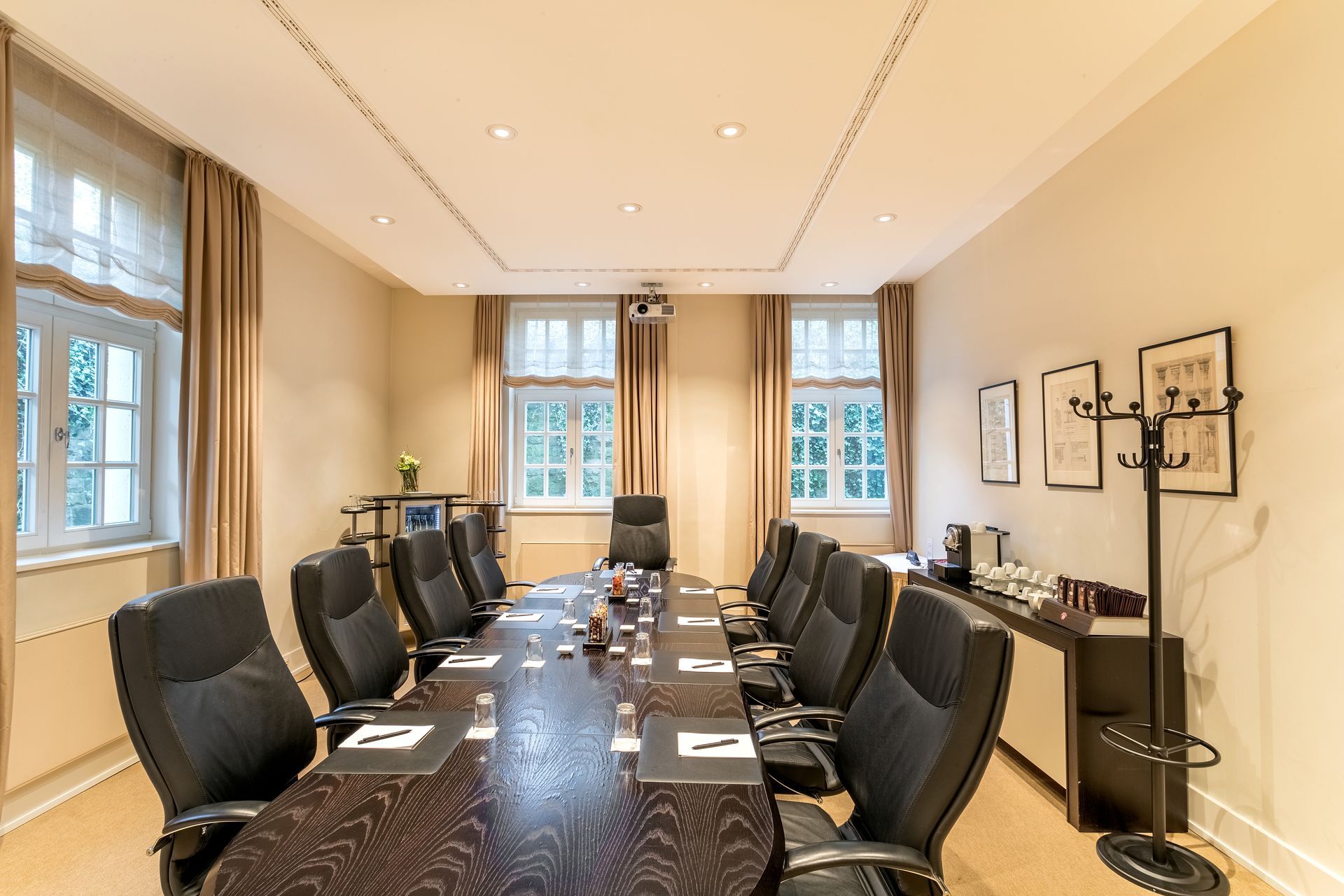 Konferenzraum mit langem Tisch, schwarzen Stühlen, Fenstern, neutralen Wänden, Kaffeestation und Garderobe.