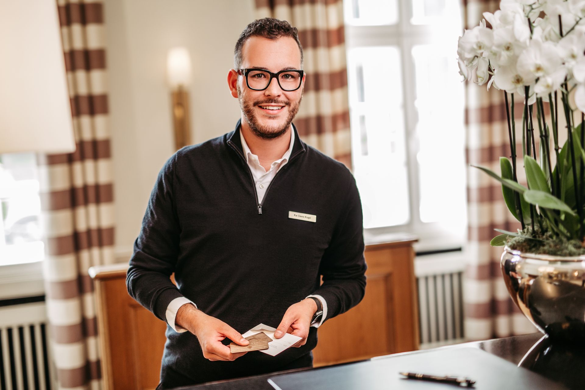 A smiling man wearing glasses and a sweater is sitting at a reception desk, holding business cards in his hand.