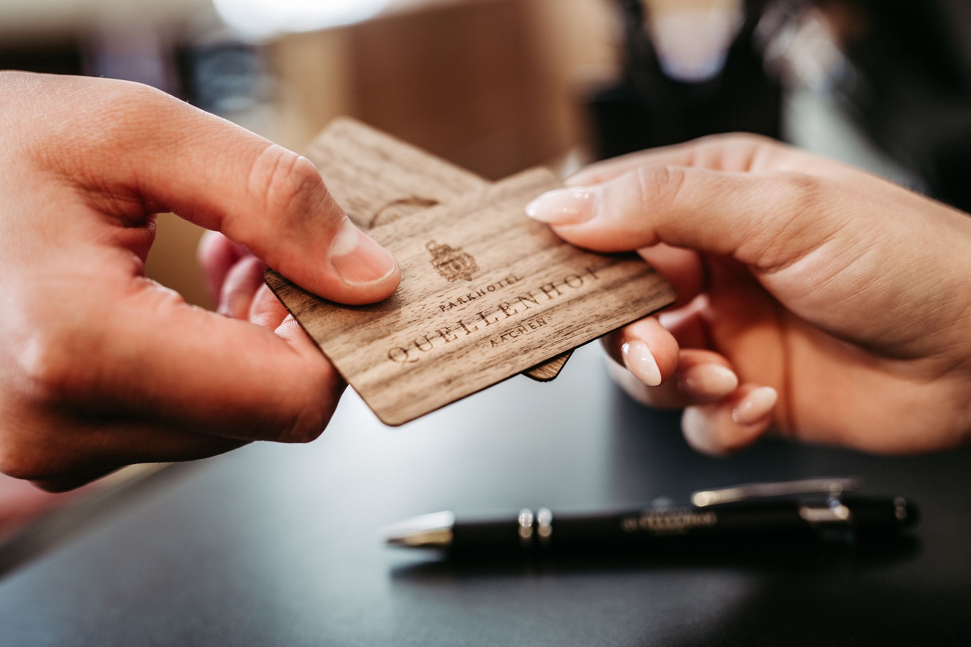 Hands are exchanging two wooden business cards across a table using a pen.