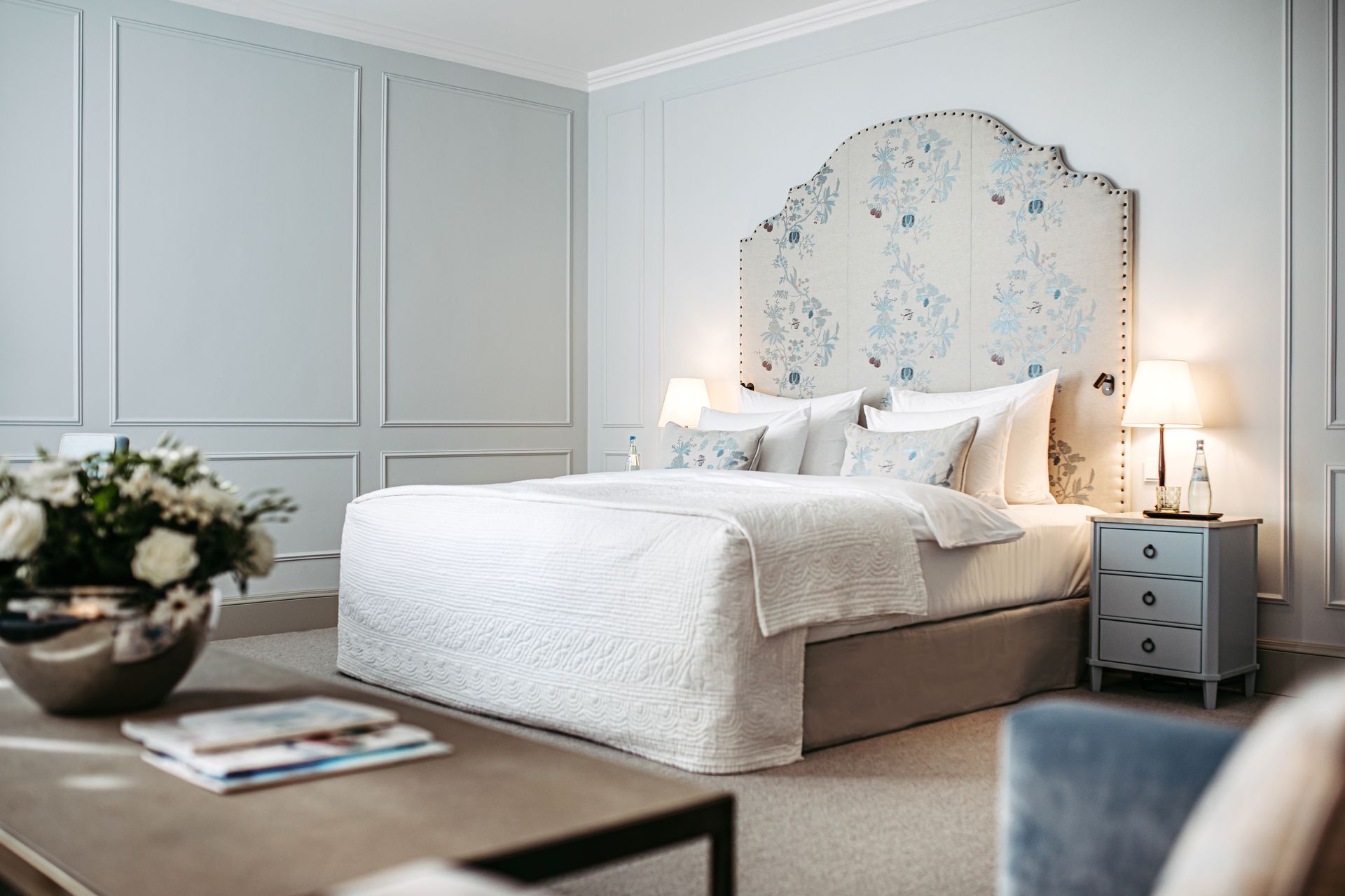A hotel room with a king-size bed featuring a floral-patterned headboard, light gray walls, and a side table with a lamp.