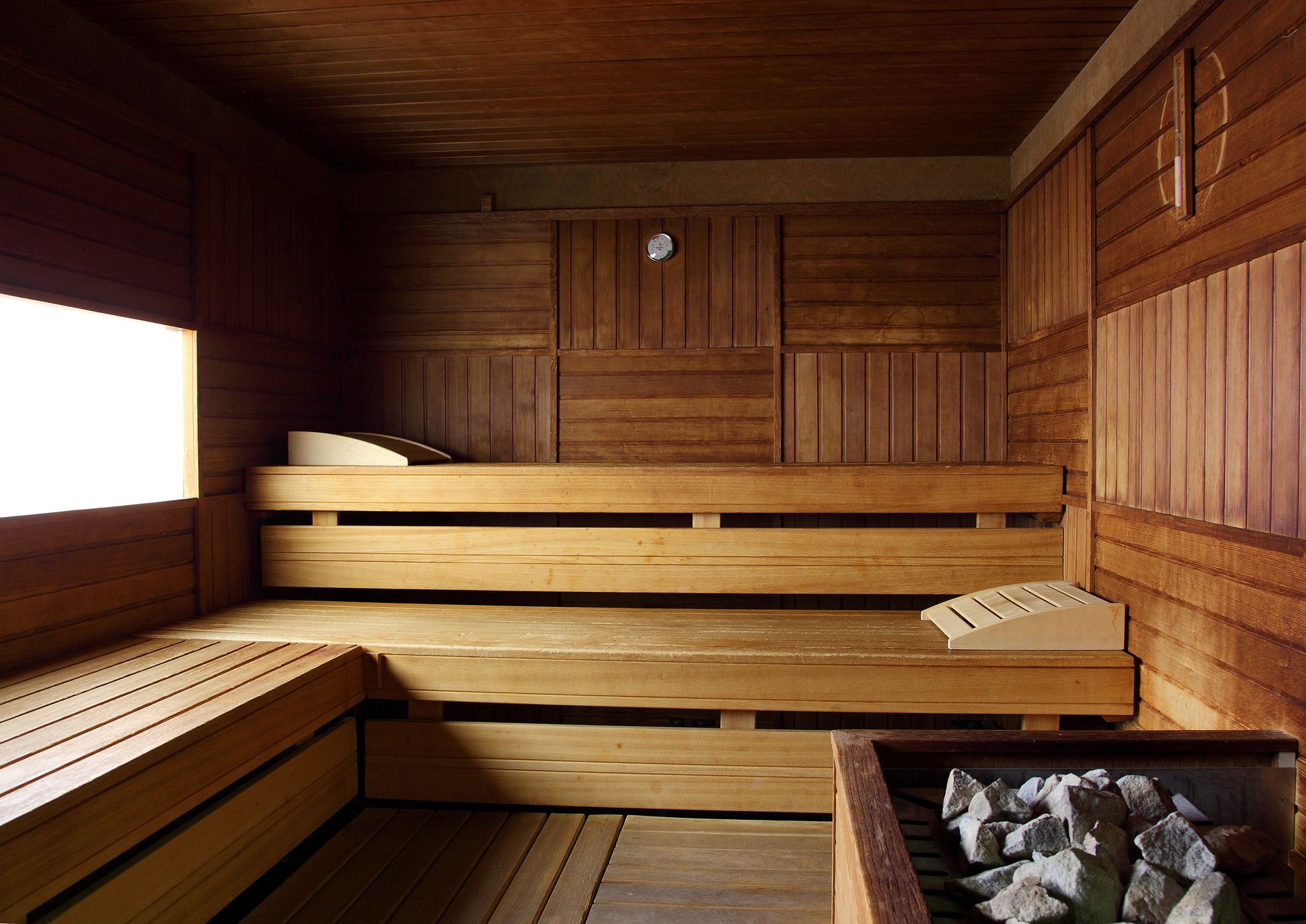 Saunainterieur aus Holz mit zweistufigen Bänken und heißen Steinen, beleuchtet durch ein Fenster.