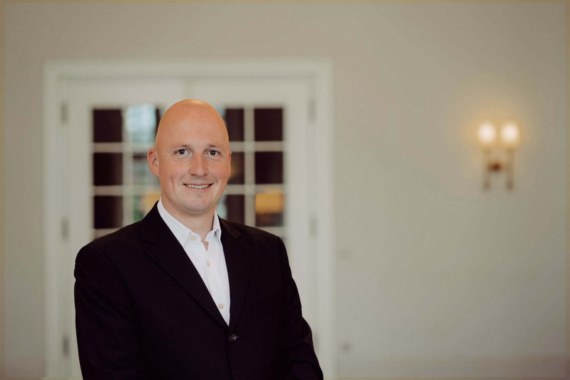Ein Mann im Anzug lächelt, er steht in der Nähe einer weißen Tür, in einem Innenraum vor neutralem Hintergrund.