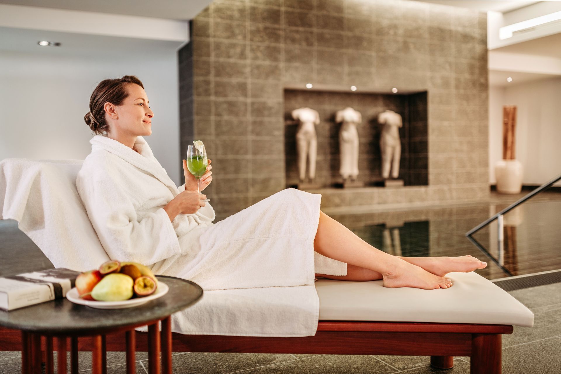 A woman in a white bathrobe is relaxing with a drink and some fruit by the pool at a spa.