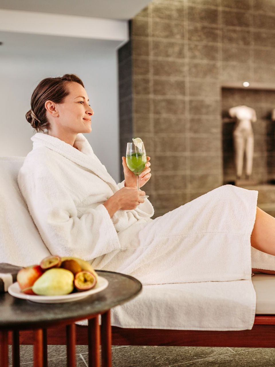 Frau entspannt im Bademantel, hält Getränk und Obstteller in der Hand, ruhige Umgebung.