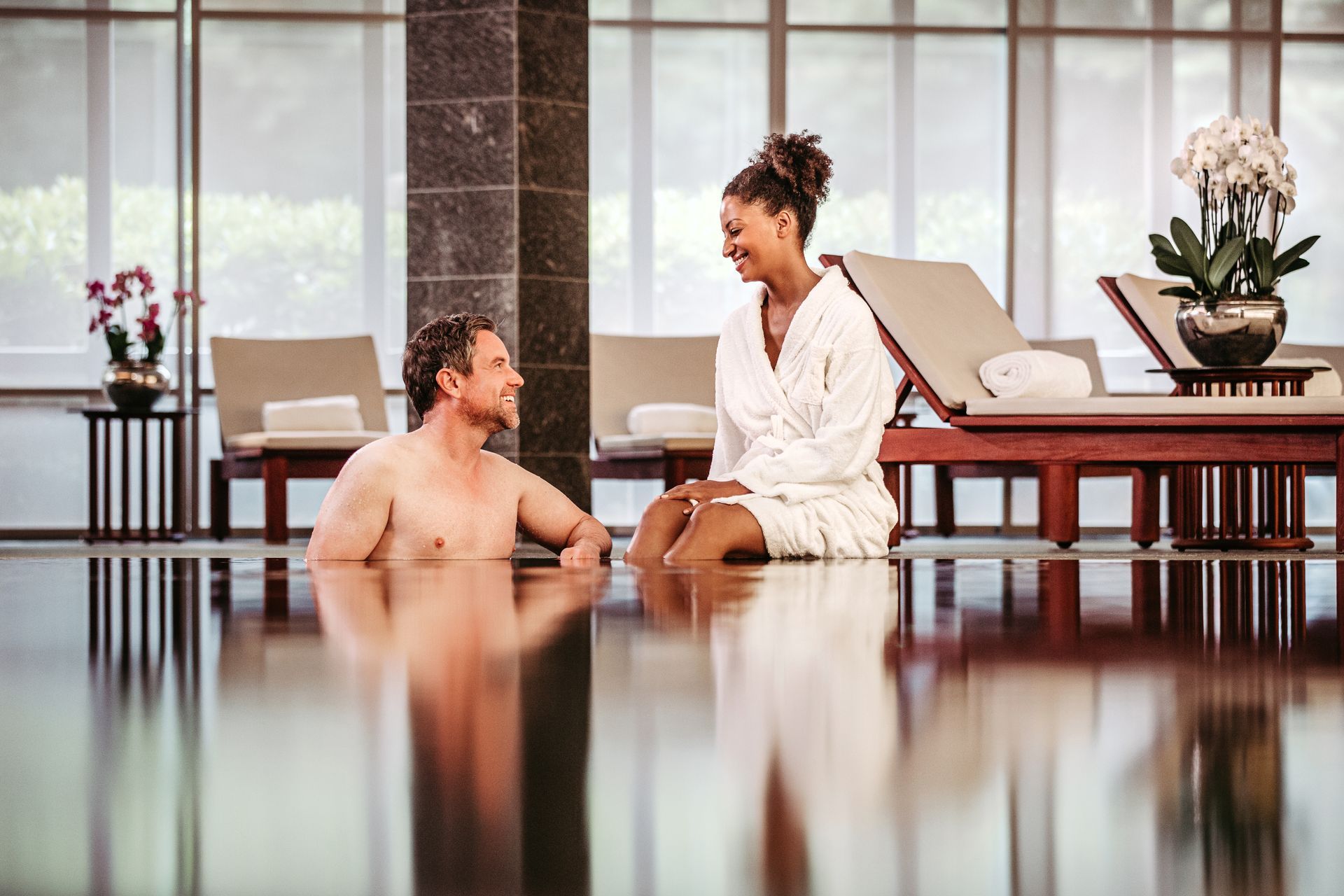 Ein Mann unterhält sich im Pool mit einer Person im Bademantel in einem Spa mit Liegestühlen und Orchideen.