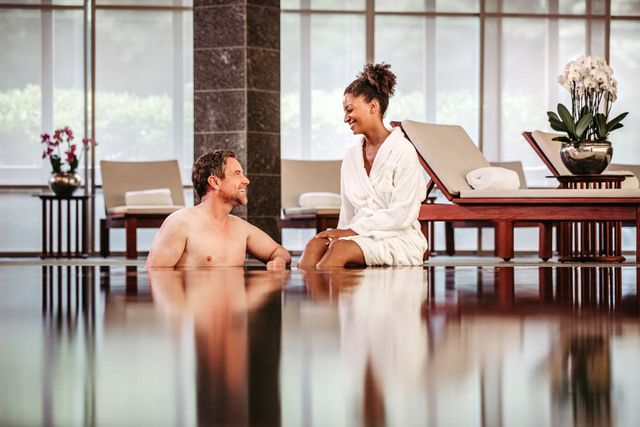 Ein Mann im Pool schaut eine Frau im Bademantel an, die lächelt; sie befinden sich in einem Spa-Bereich.