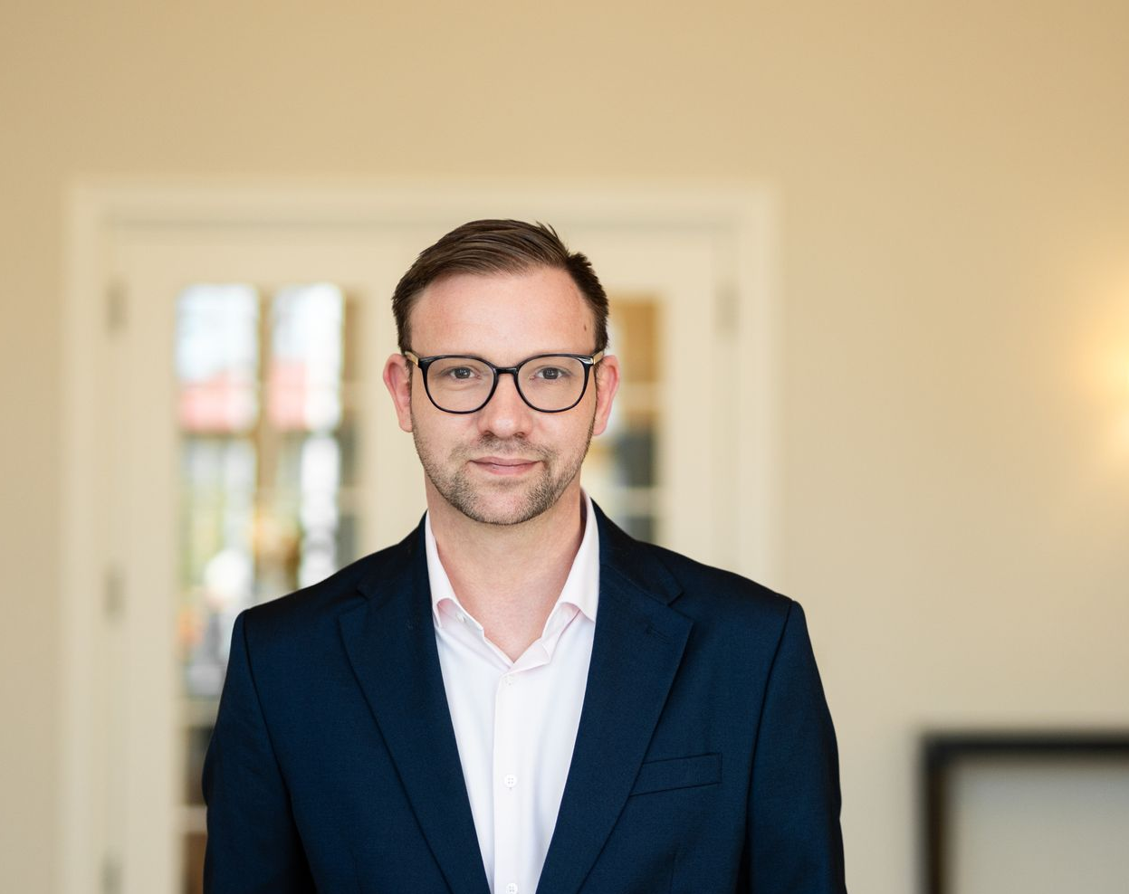 Eine lächelnde Frau mit blonden Haaren, die einen schwarzen Blazer und ein weißes Hemd trägt, steht vor einer weißen Tür.