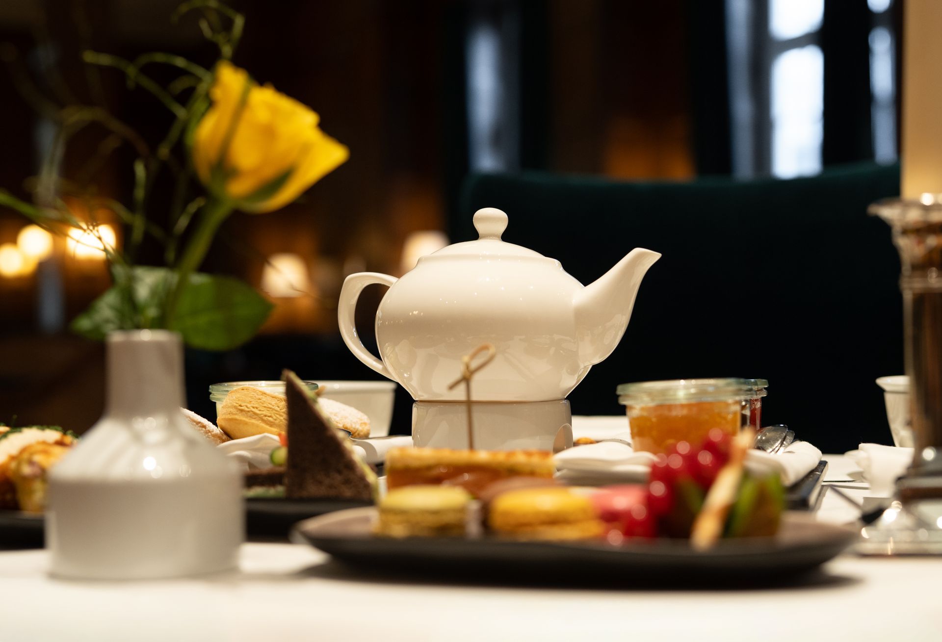 Un service à thé et des coupes à champagne sur une nappe blanche. Une étagère à pâtisseries. Une pièce faiblement éclairée.