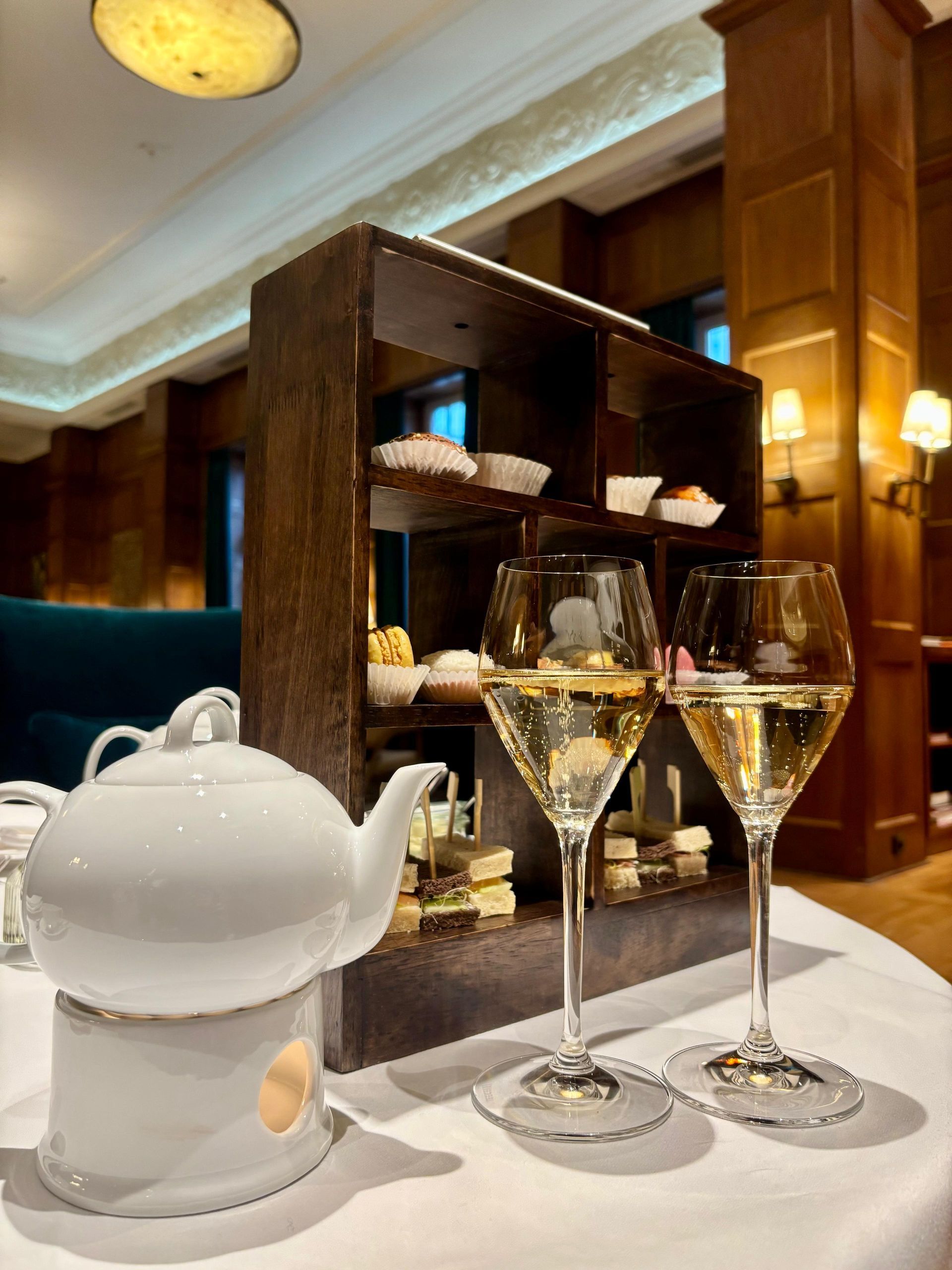 Teeservice und Champagnergläser auf einer weißen Tischdecke. Eine Etagere mit Gebäck. Schwach beleuchteter Raum mit Holzvertäfelung.