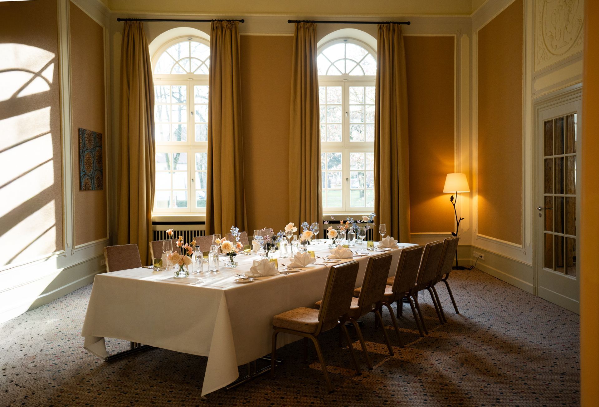 Elegantes Esszimmer mit langem, für ein Essen gedecktem Tisch, hohem Blumengesteck in der Mitte und großen Fenstern.