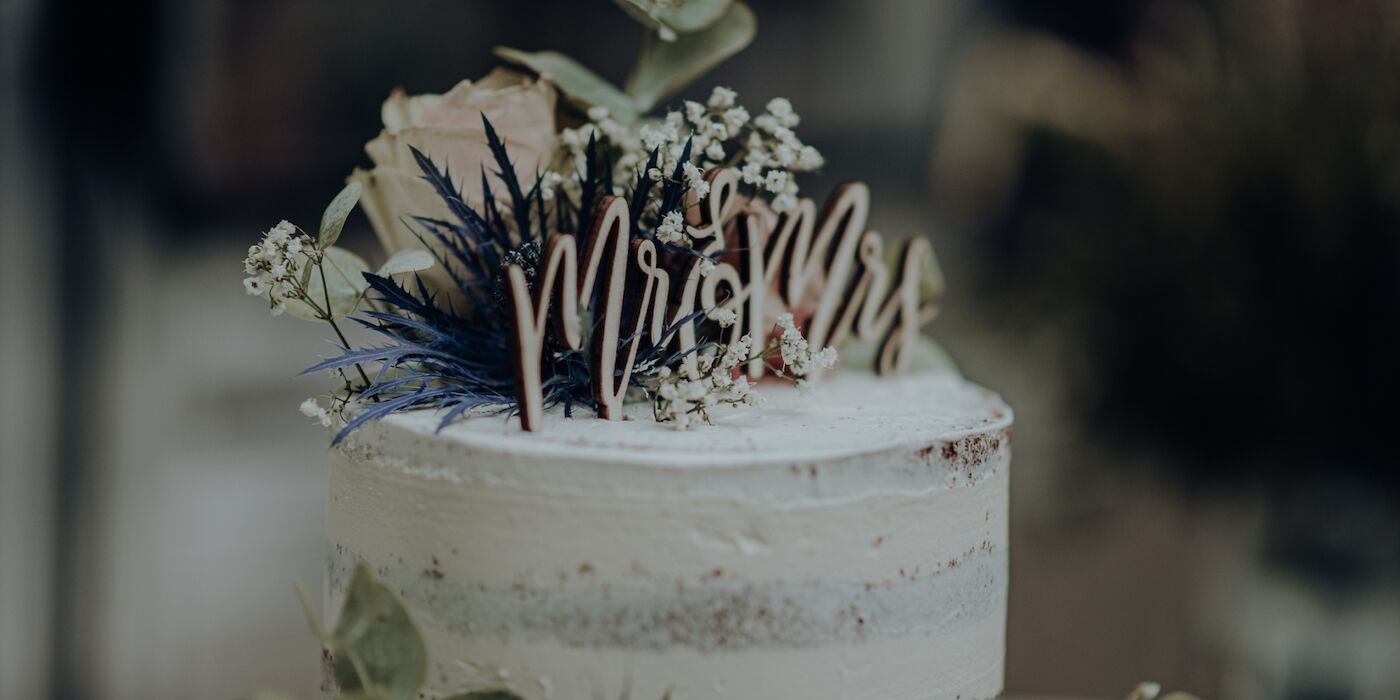 Hochzeitstorte mit der Aufschrift „Mrs.“ als Tortenaufsatz, verziert mit Blumen und Grünzeug.