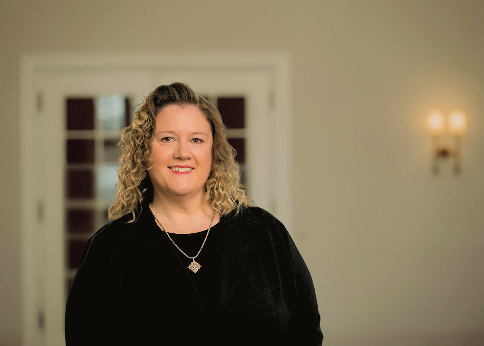 Eine Frau mit lockigem Haar lächelt. Sie trägt ein schwarzes Hemd und eine schwarze Halskette und steht in einem Gebäude.