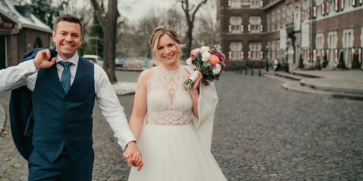 Ein frischvermähltes Paar hält Händchen, lächelt und schreitet über Kopfsteinpflaster auf den Betrachter zu. Die Frau hält einen Blumenstrauß. Im Hintergrund ist ein Gebäude zu sehen.