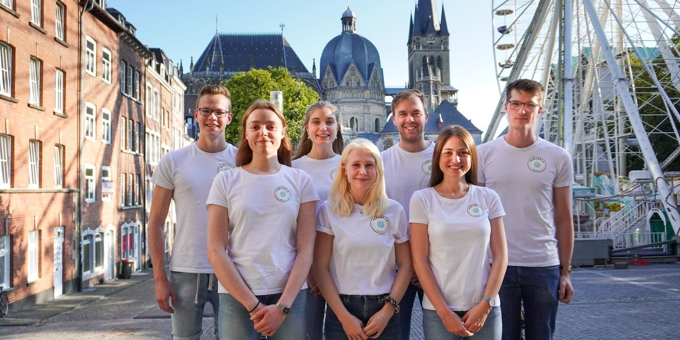 A group of people wearing white T-shirts is standing in front of some buildings and a Ferris wheel.
