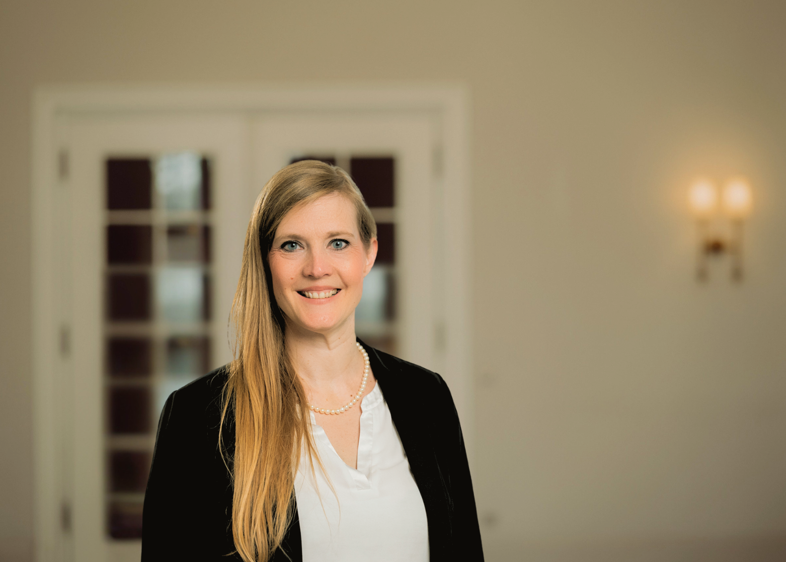 Eine lächelnde Frau mit blonden Haaren, die einen schwarzen Blazer und ein weißes Hemd trägt, steht vor einer weißen Tür.