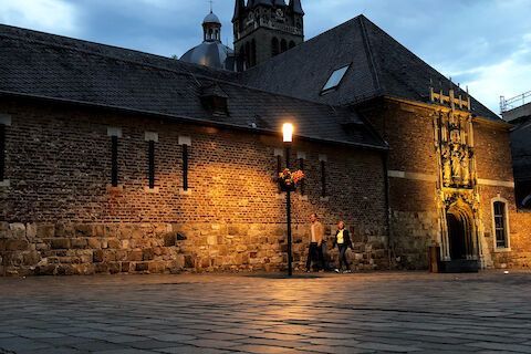 A stone facade with a lamppost, two passersby, twilight.
