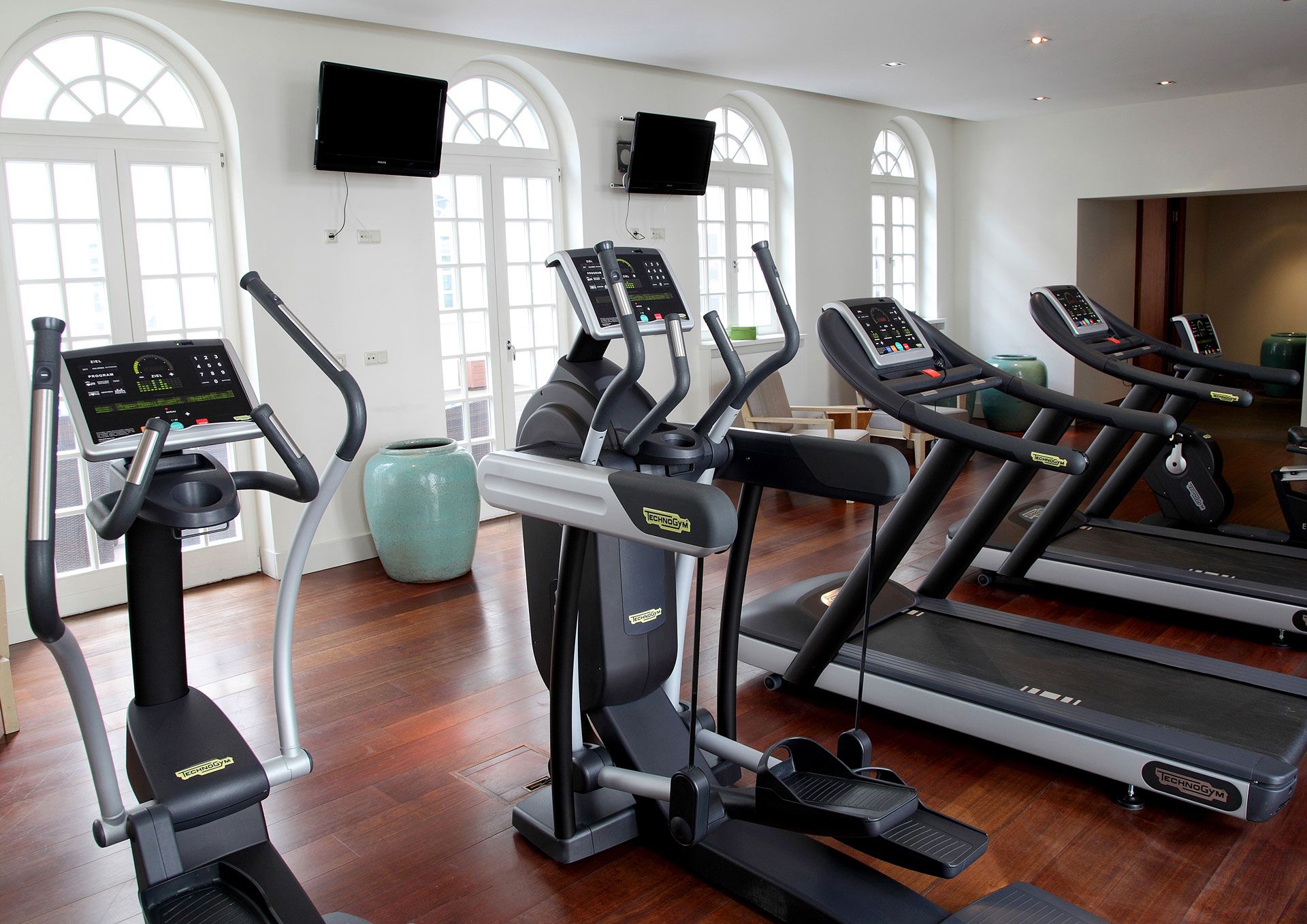 Fitnessstudio-Interieur mit Ellipsentrainern, Laufbändern und Fernsehern; Bogenfenster und Holzboden.