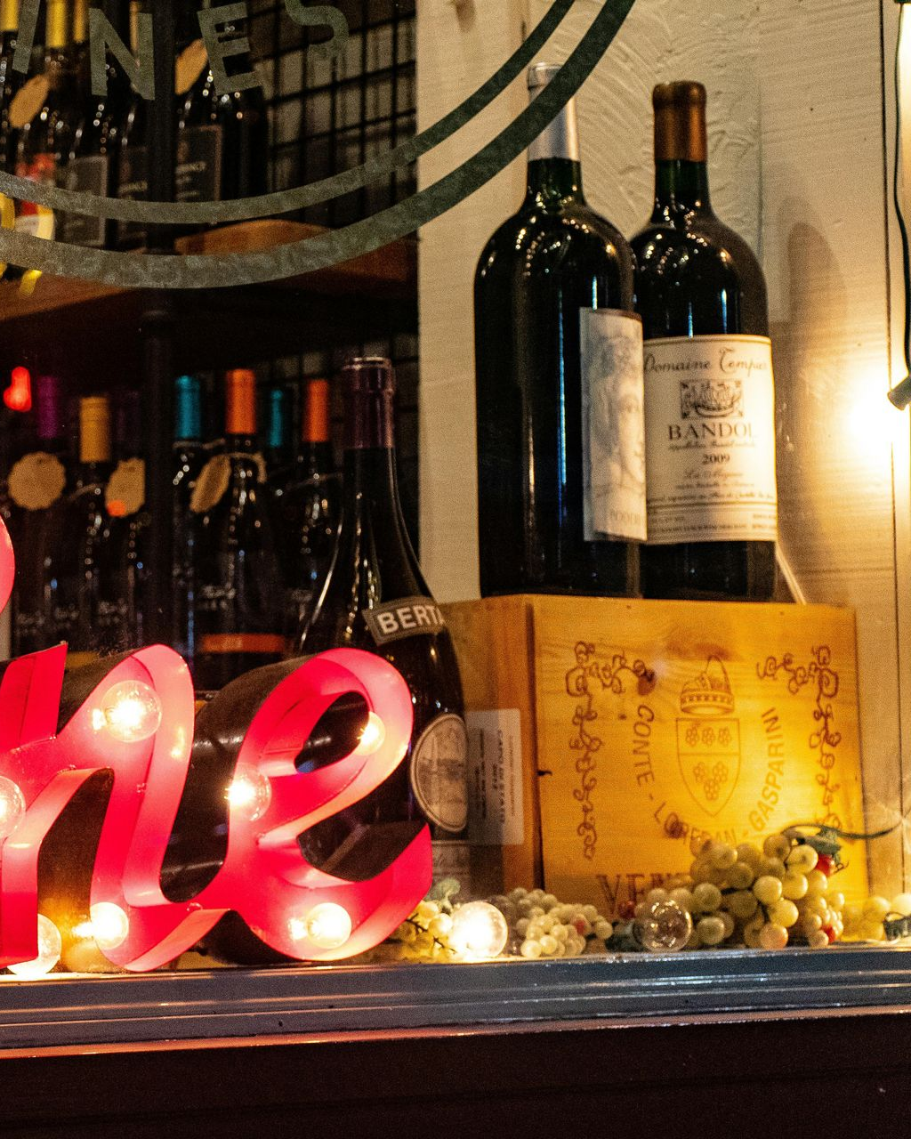 Weinflaschen sind in einem Schaufenster ausgestellt, zusammen mit einem leuchtenden „Wein“-Schild und Trauben.