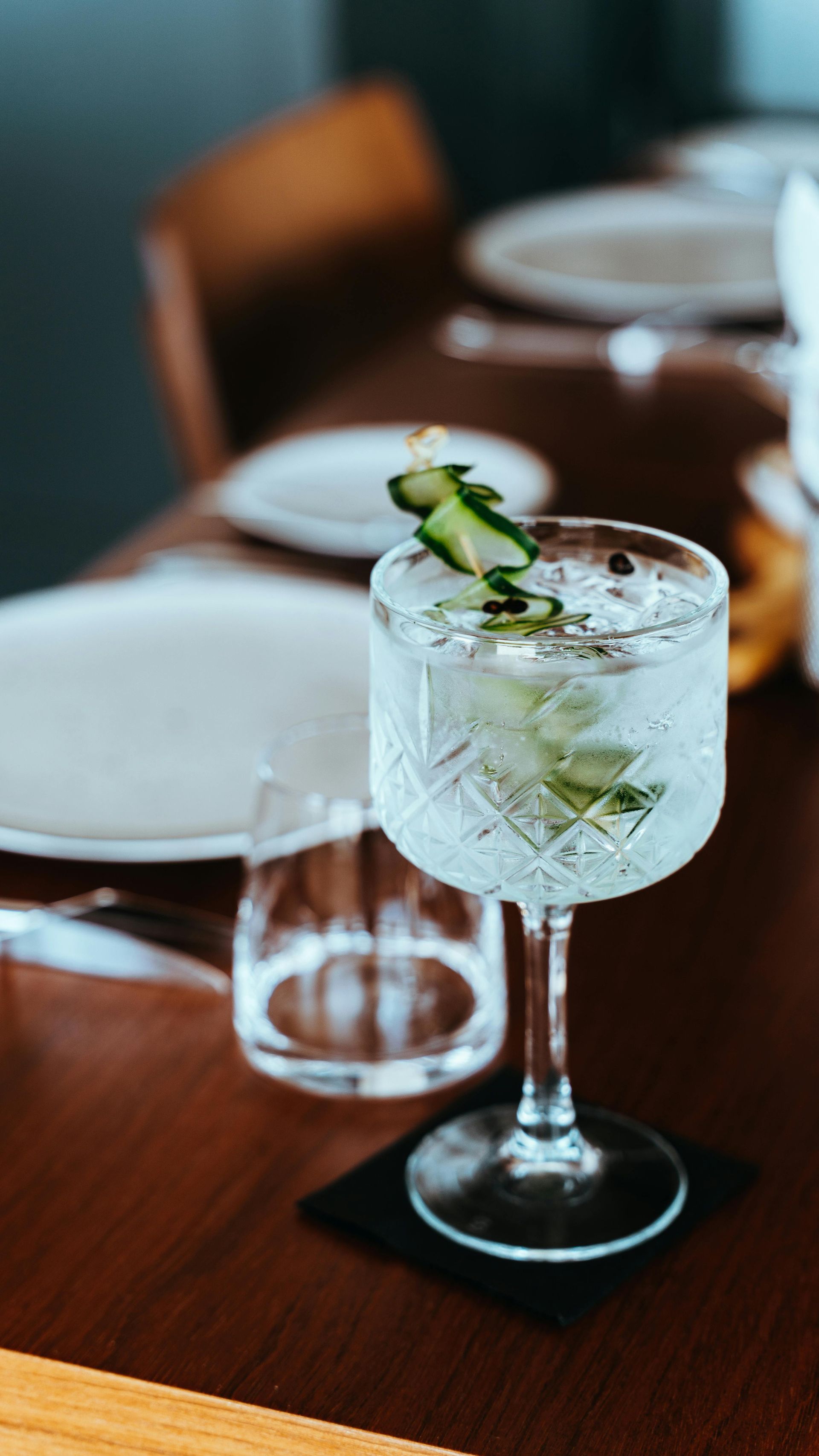 A cocktail in a tall glass garnished with cucumber, sitting on a dark wooden table next to a water glass and plates.