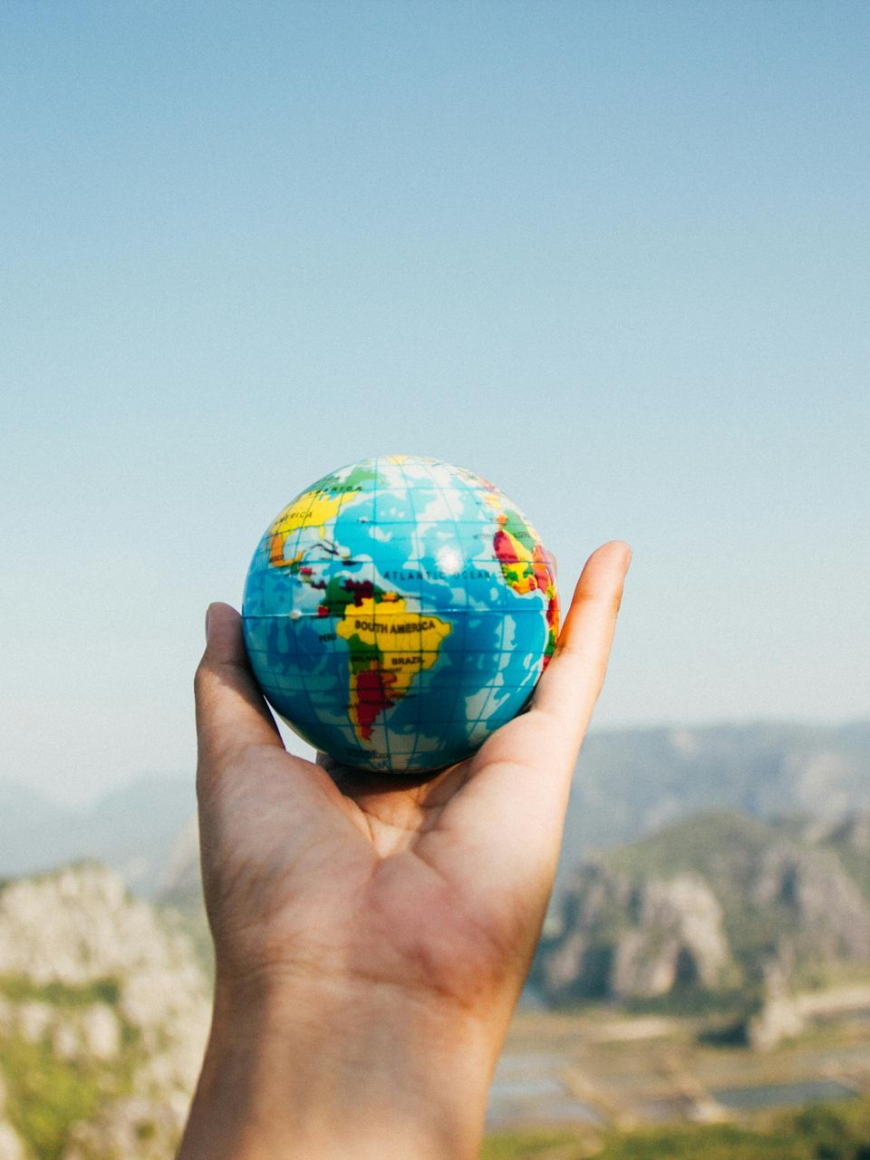 A hand is holding a small globe against a blurred backdrop of mountains and sky.