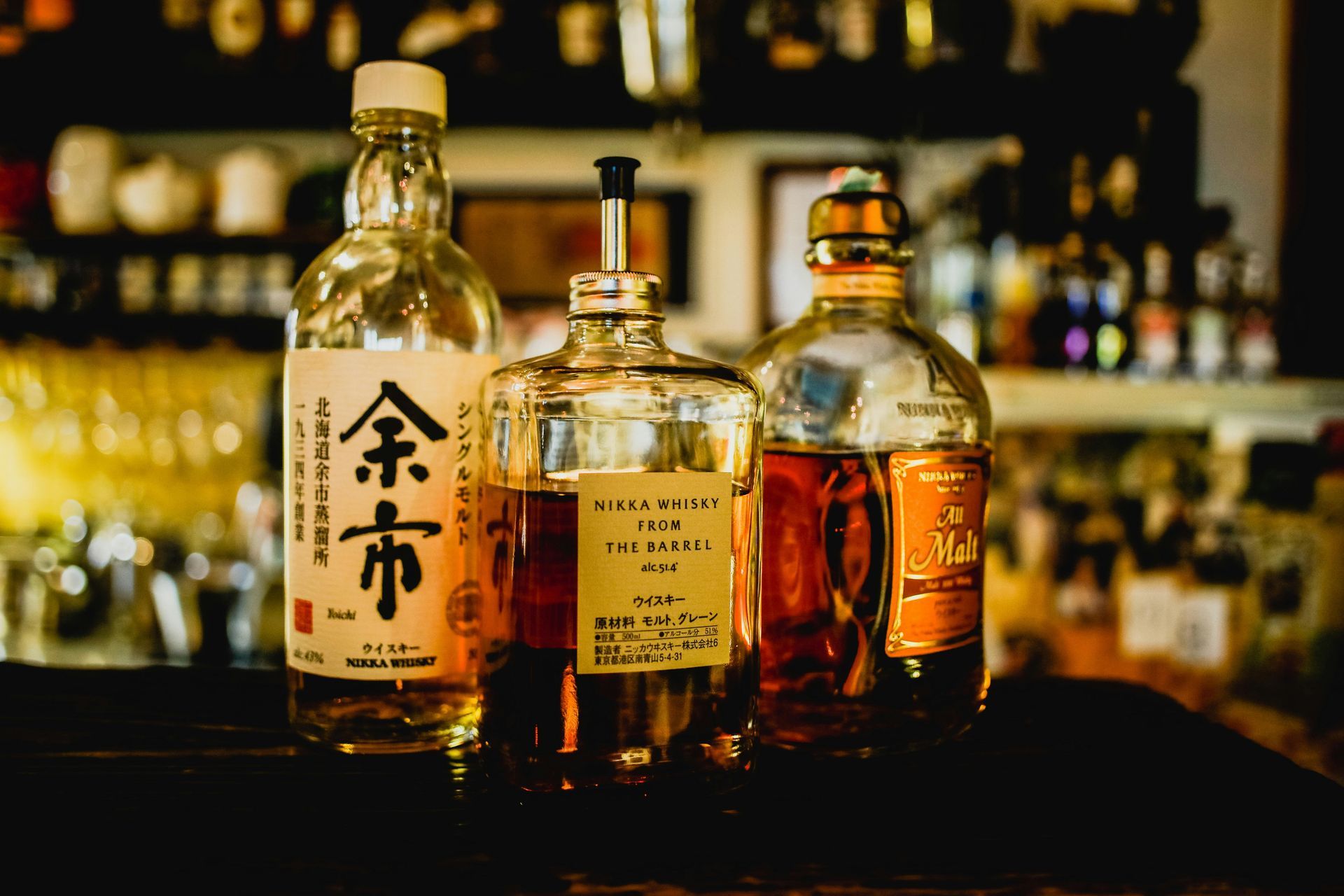 Three bottles of whiskey on a bar, bathed in amber light, against a blurred backdrop of bar shelves.