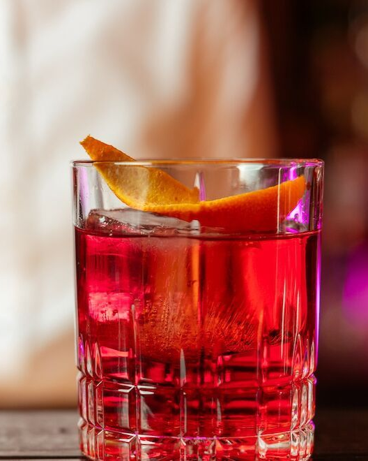 A red cocktail in a crystal glass, garnished with an orange peel.