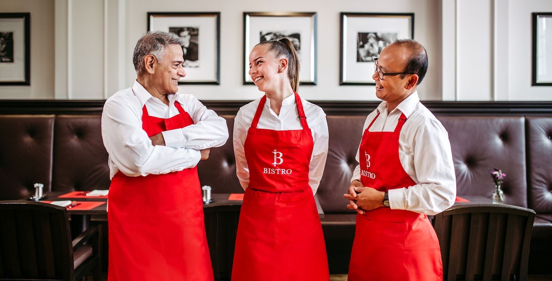 Three people wearing red aprons and white shirts, smiling in a restaurant.