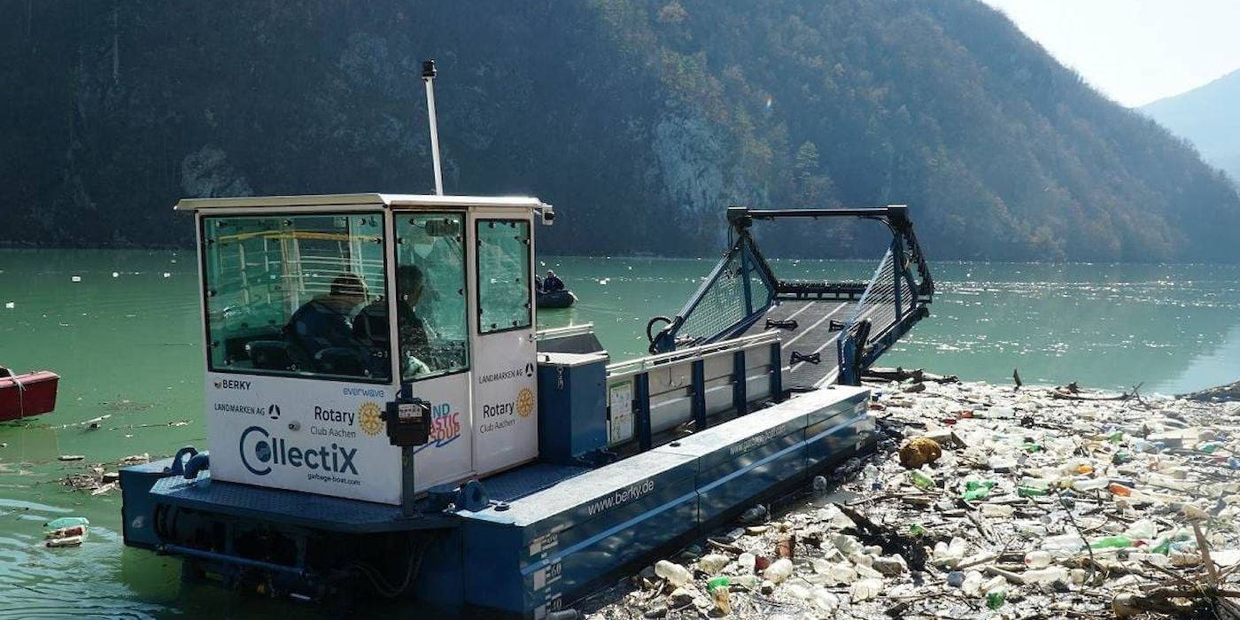 A garbage collection boat navigates across a polluted lake, with mountains rising in the background.