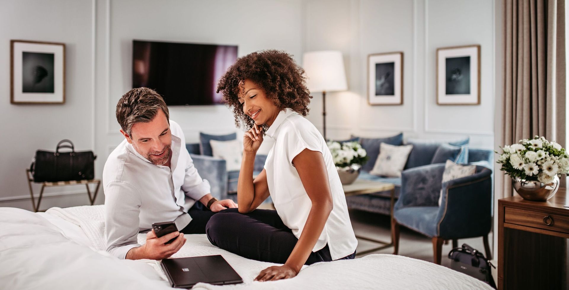 Ein Paar sitzt auf einem Bett und schaut auf ein Handy; das Zimmer ist mit einem Sofa und Kunstwerken ausgestattet.