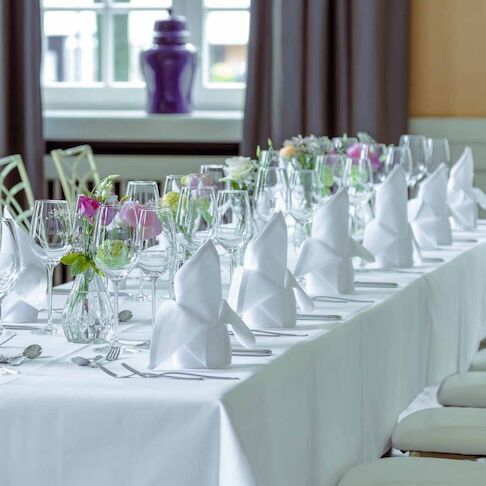 Eine lange Tafel, gedeckt für einen formellen Anlass mit weißer Tischdecke, Gläsern, Servietten und Blumenarrangements.