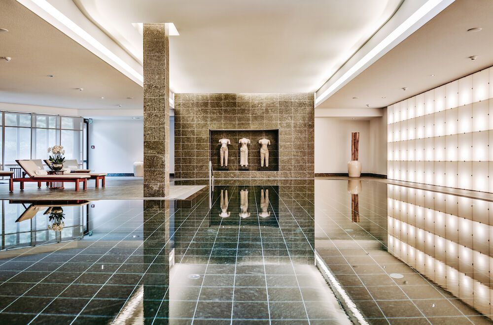 An indoor pool with a dark tiled floor that reflects the sculptures, architecture, and atmospheric lighting.