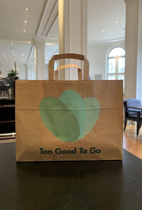 Eine braune Papiertüte mit grünem Logo und dem Aufdruck „Too Good To Go“ liegt auf einem Tisch.
