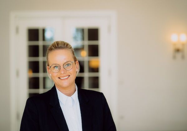 A woman in a black blazer and glasses is smiling, standing next to white double doors.