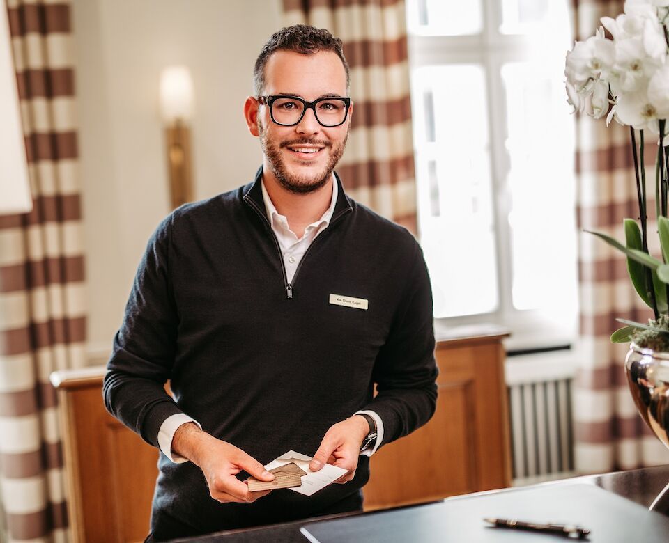 A smiling man wearing glasses, holding cards in his hand, is standing behind a hotel reception desk.